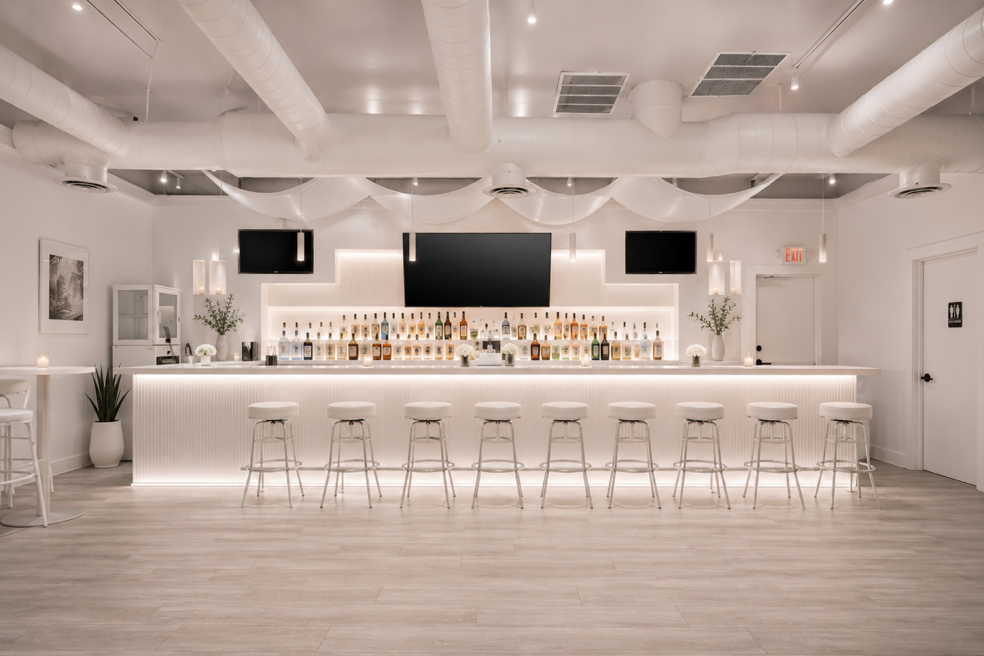 A long illuminated bar in white with a backlit bottle display and a row of stools